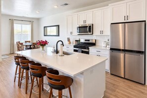 A modern kitchen with white cabinetry, stainless steel appliances, a central island with bar stools, and a dining area with a table and chairs. - Spacious Retreat w/ Pool, Pickleball & Patio in UT (Washington)