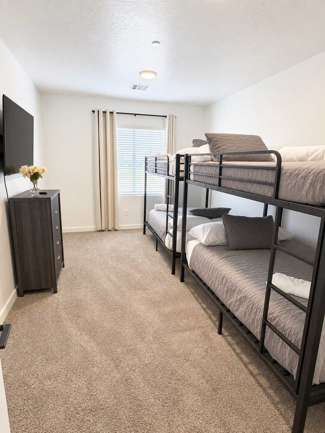 Een slaapkamer met een stapelbed, een flatscreen televisie op een kast, een raam met gordijnen en een vloerkleed.