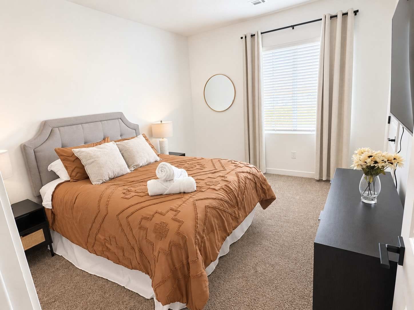 A hotel room with a bed, a bedside table, a lamp, a round mirror, a window with curtains, and a vase with flowers.