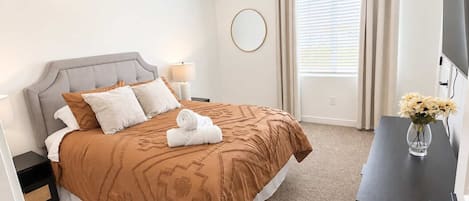A hotel room with a bed, a bedside table, a lamp, a round mirror, a window with curtains, and a vase with flowers.