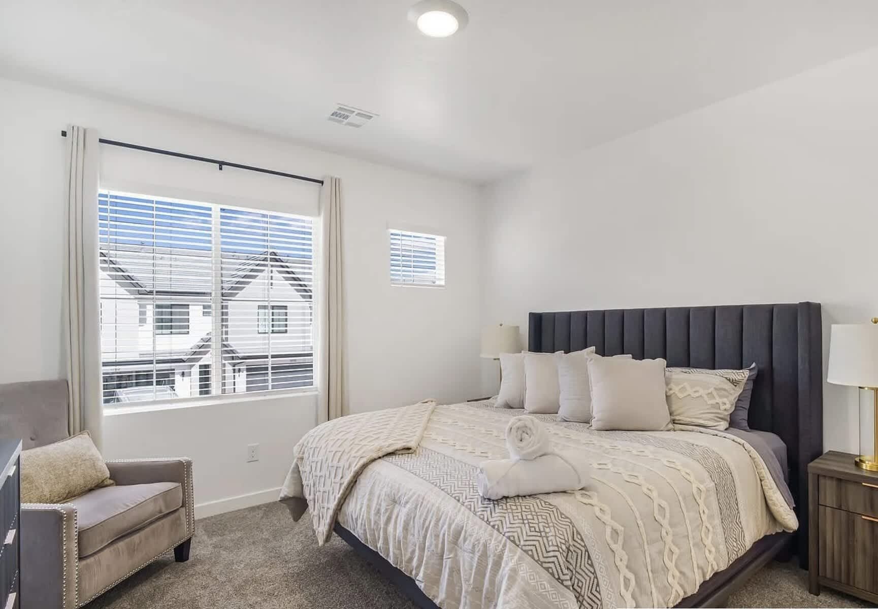 Quarto com cama, cadeira, mesa de cabeceira e janela com persianas.