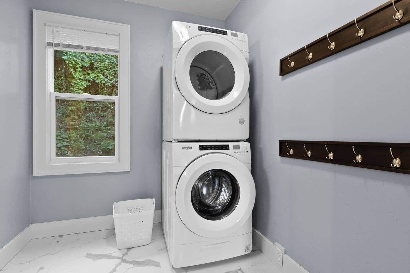 A laundry room with two stacked front-loading washing machines, a window with a view of greenery, and a coat rack with hooks.