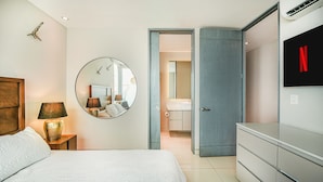 Quarto com cama, abajur, espelho redondo e TV fixada na parede.