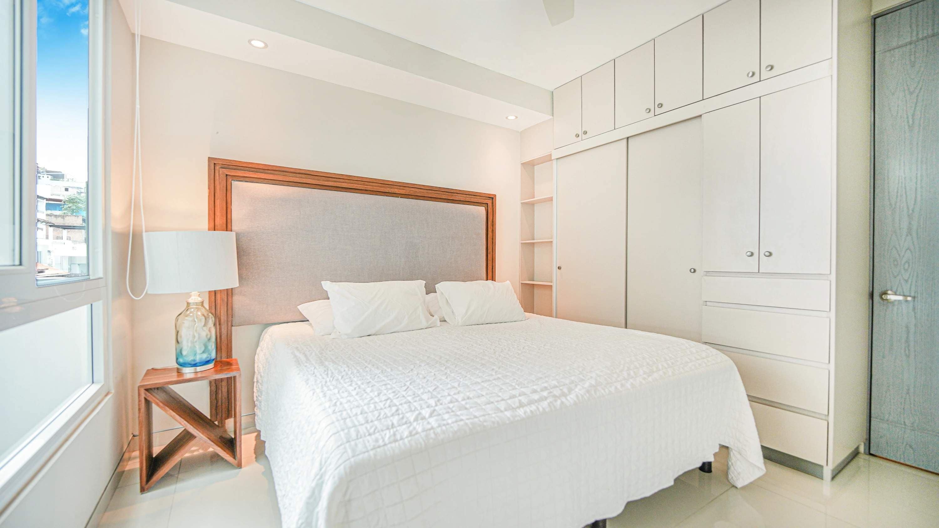 Een hotelkamer met een bed, nachtkastje, lamp, kledingkast en uitzicht op buiten.