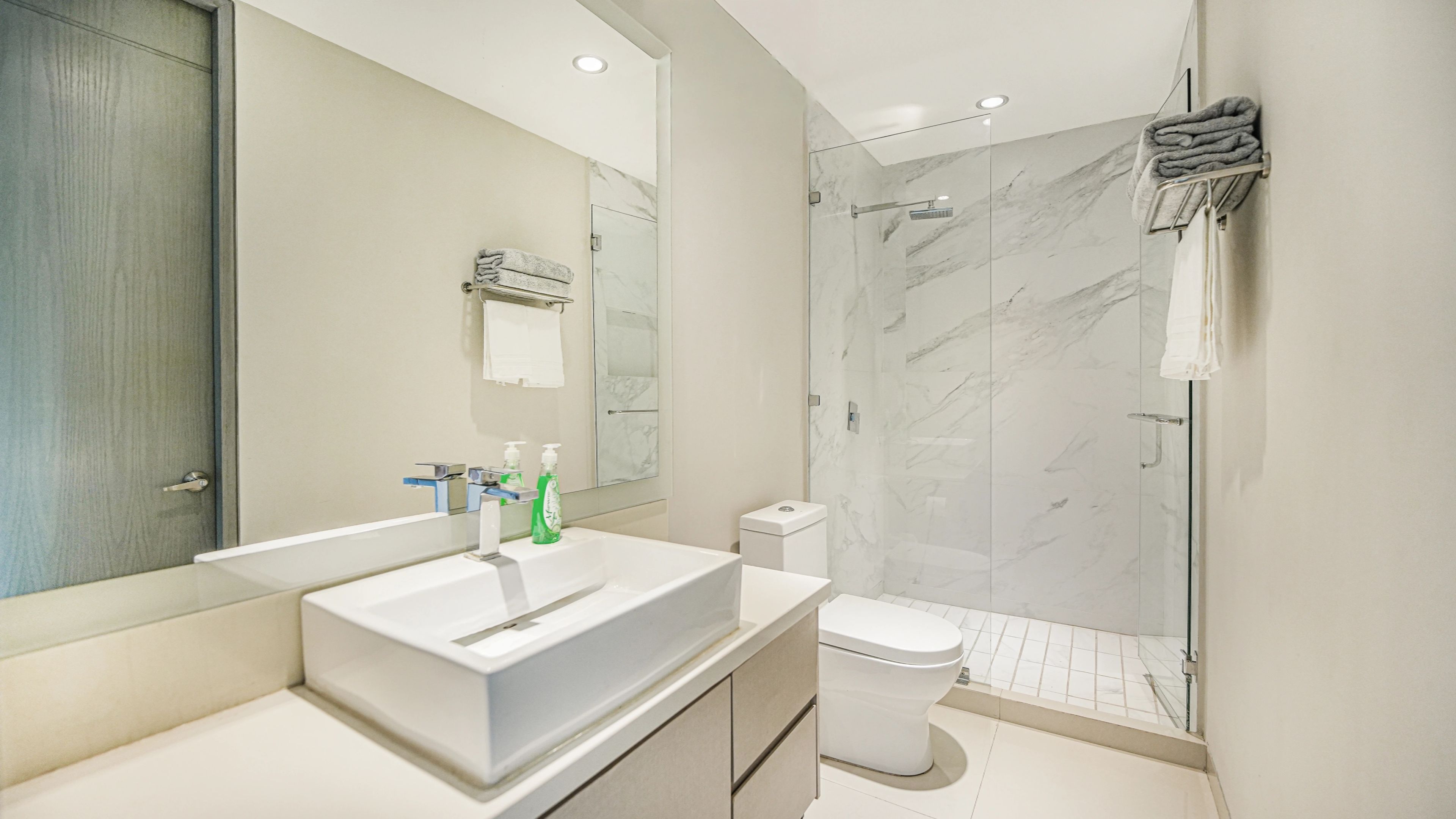 Un baño moderno con un espejo grande, un lavamanos blanco, un inodoro y una ducha con mampara de cristal.