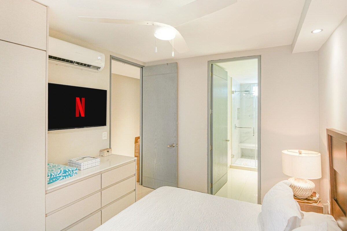 Habitación de hotel con cama, televisión montada en la pared, armario, mesita de noche con lámpara y puerta de vidrio que da a la zona de ducha.