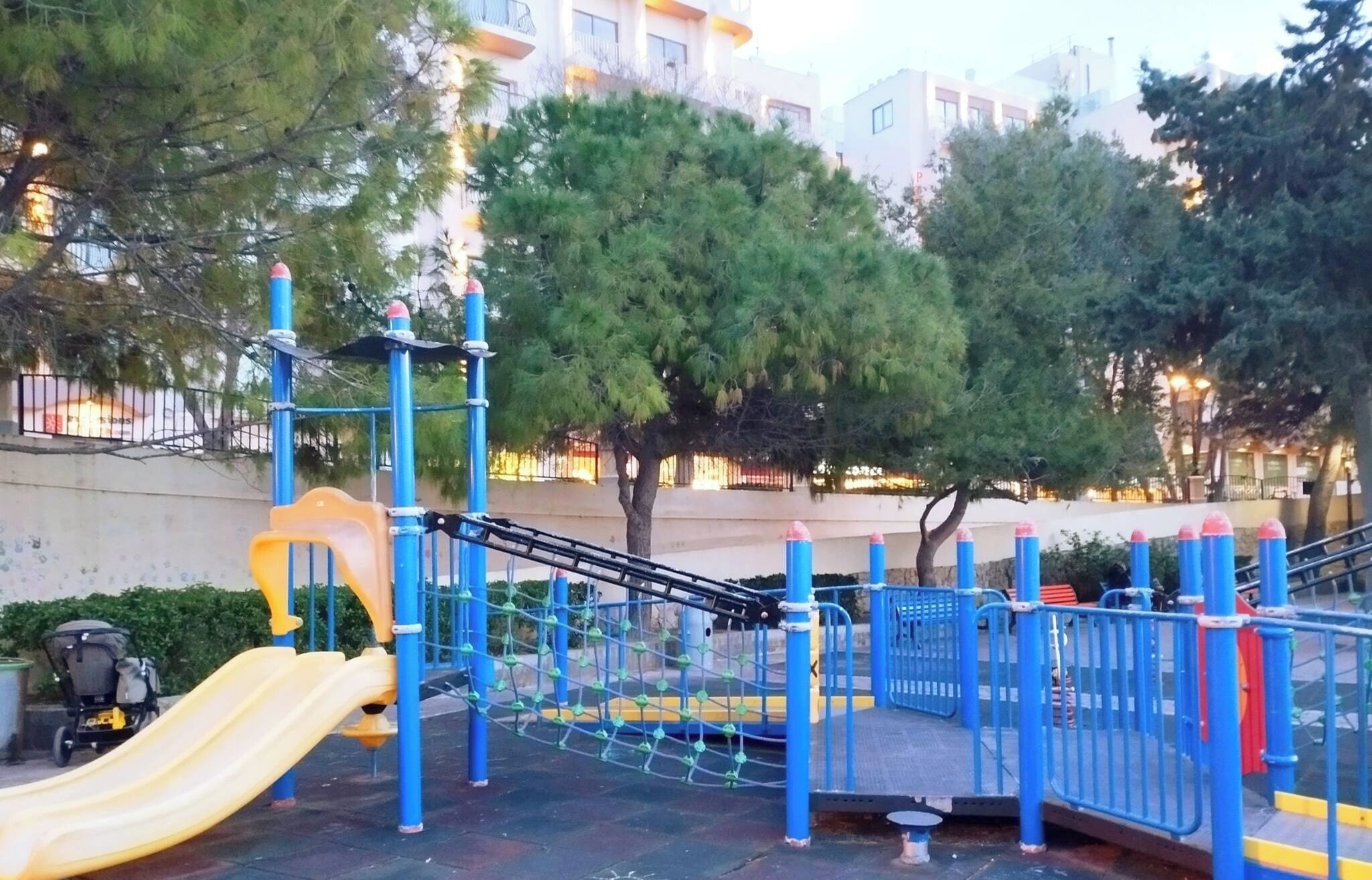 A playground with a slide and climbing structures, surrounded by trees and buildings.