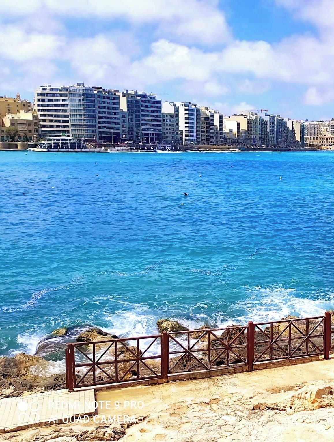 A coastal cityscape with buildings along the waterfront, a rocky shoreline, and a metal railing in the foreground.
