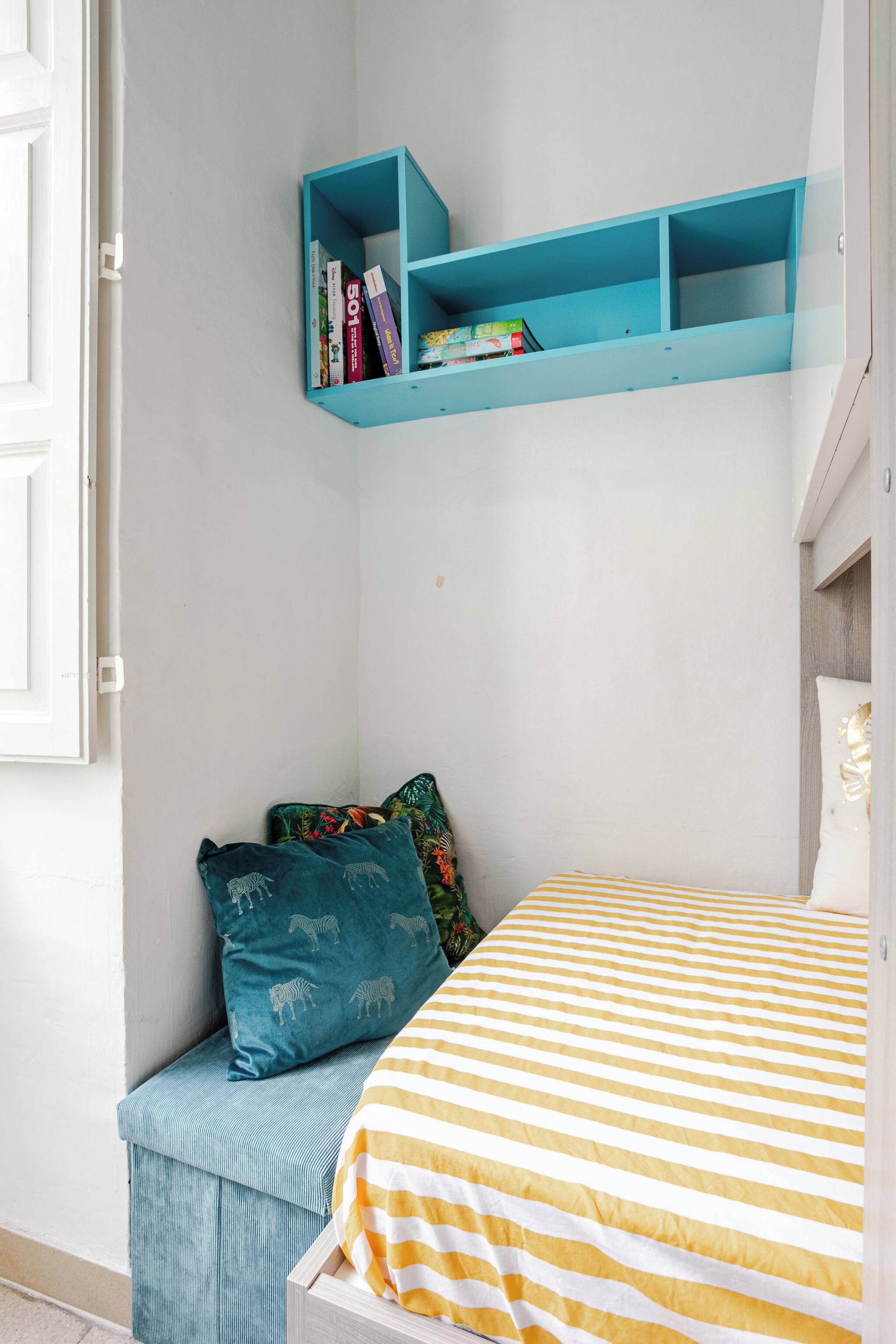 Un dormitorio con una cama, una estantería azul con libros, una ventana y una puerta.