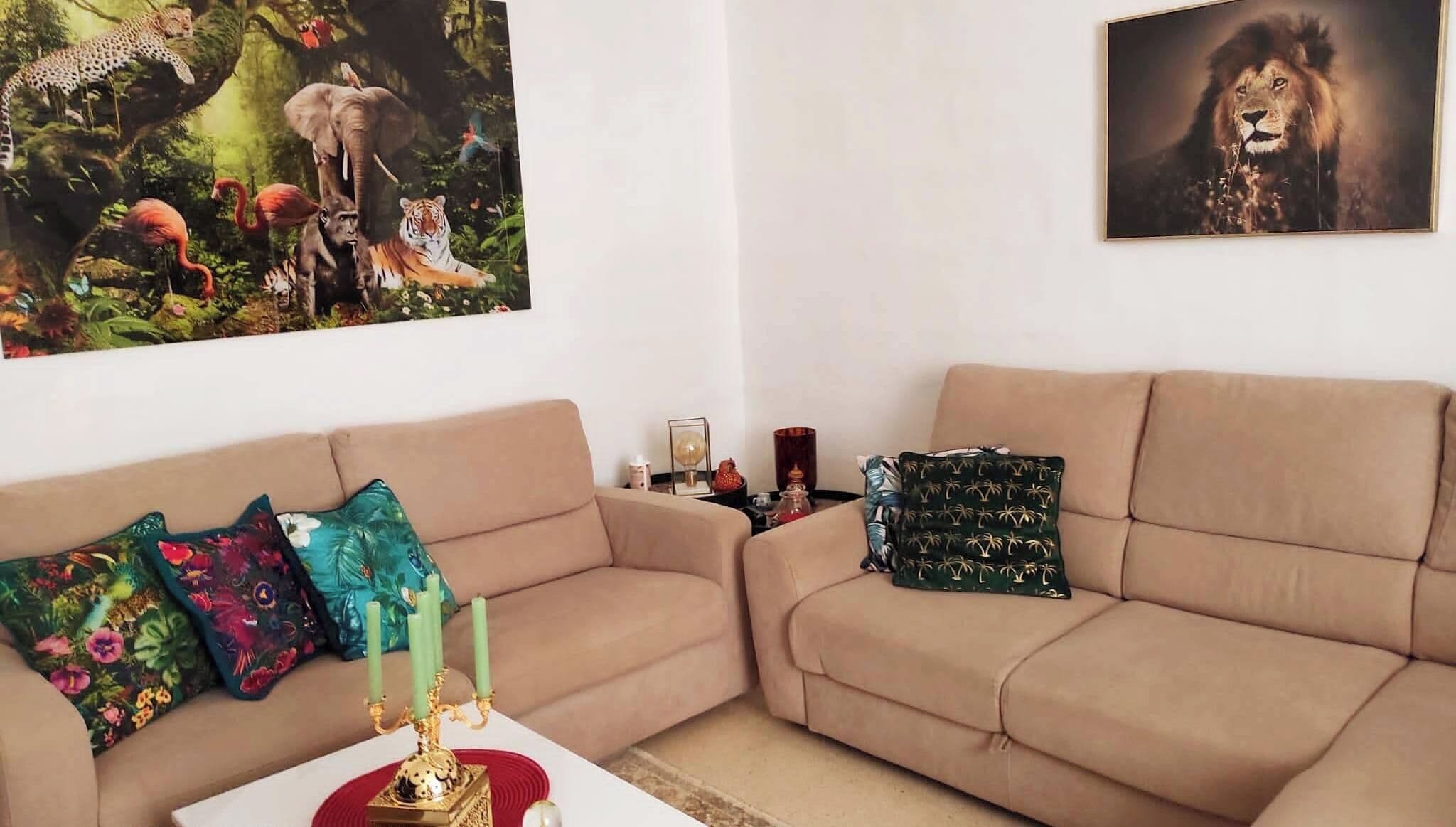 Un salón con un sofá color beige, cojines de colores, un candelabro dorado y dos cuadros enmarcados con motivos de fauna.