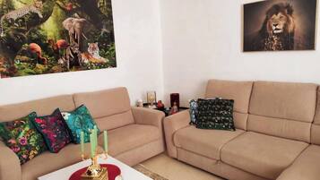 Un salón con un sofá color beige, cojines de colores, un candelabro dorado y dos cuadros enmarcados con motivos de fauna.