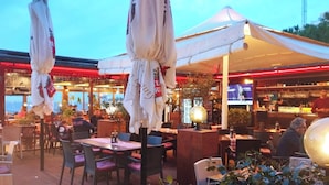 Et udendørs spiseområde med borde, stole, parasoller og en bar med et tv.