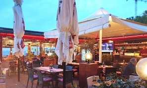 Un'area esterna con tavoli e sedie, ombrelloni e un angolo bar con schermo televisivo.