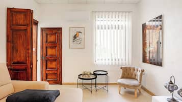 A living room with a sofa, a chair, two small tables, a large window with blinds, and a wall-mounted air conditioning unit.