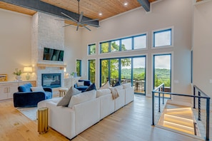 A spacious living area with a large sofa, a fireplace, and multiple windows offering a view of the outdoors. - SkySong Cottage | 4BR Mountain View Highlands (Macon County)