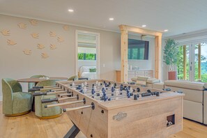 A modern living room with a foosball table, a sofa, a coffee table, and a large window with a view of greenery. - SkySong Cottage | 4BR Mountain View Highlands (Macon County)