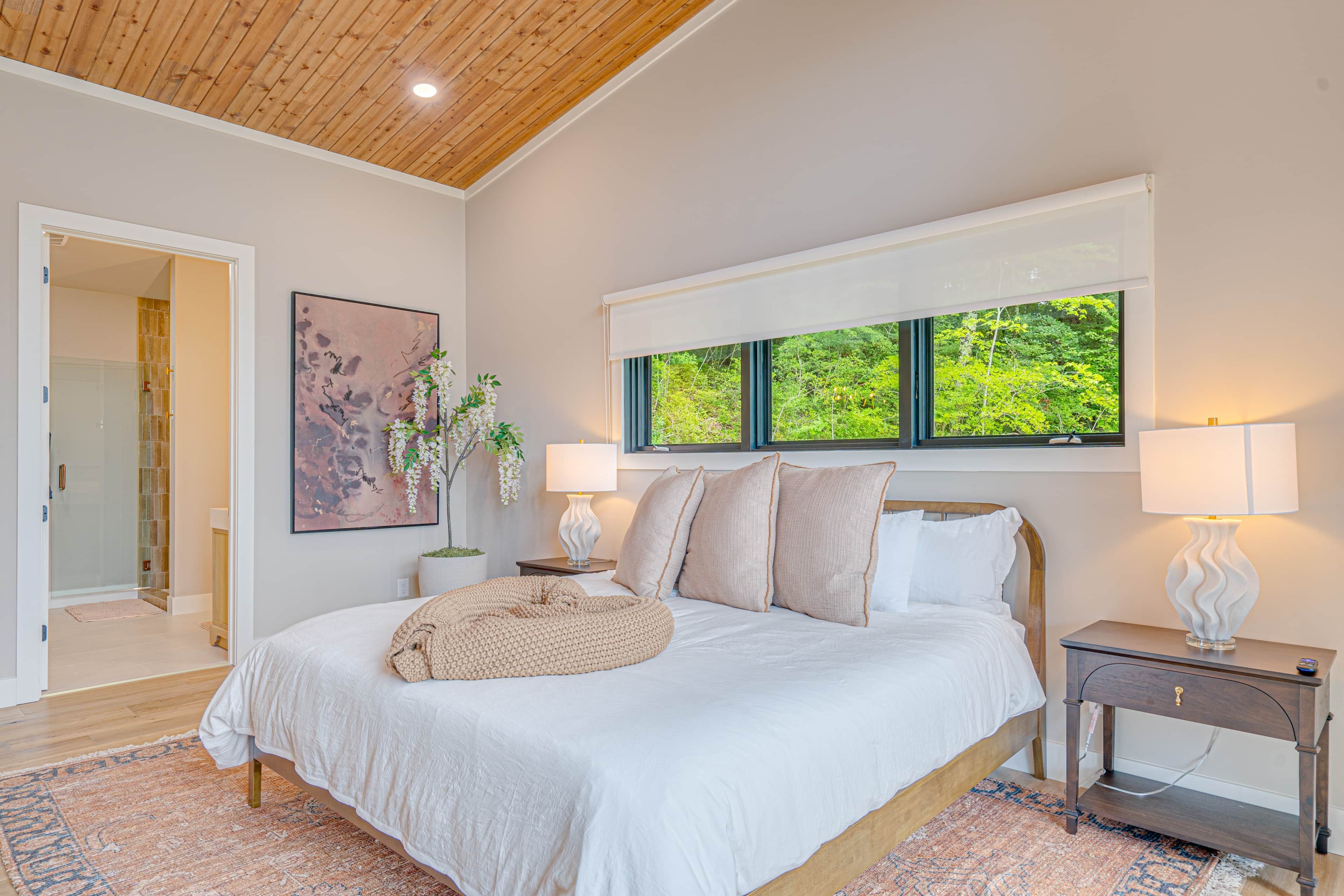 A bedroom with a large bed, a window with a view of greenery, a bedside table with a lamp, and a framed artwork on the wall.