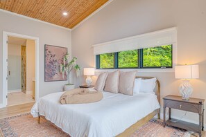 Quarto com cama grande, janela com vista para vegetação, mesa de cabeceira com abajur e quadro na parede.