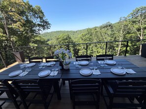 Área de refeições ao ar livre com mesas para quatro pessoas, rodeada por um deck de madeira e com vista para uma encosta arborizada.