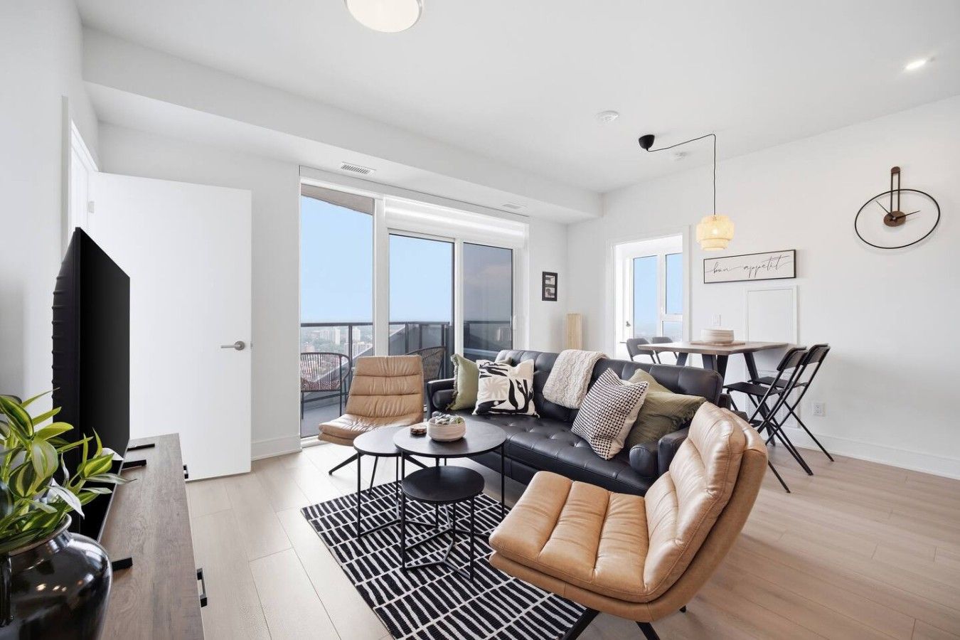 A modern living room with a black leather sofa, a beige armchair, a round coffee table, and a dining area with a table and chairs.