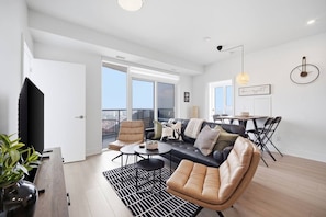 Ein modernes Wohnzimmer mit einem schwarzen Ledersofa, einem beigefarbenen Sessel, einem runden Couchtisch und einem Essbereich mit Tisch und Stühlen.