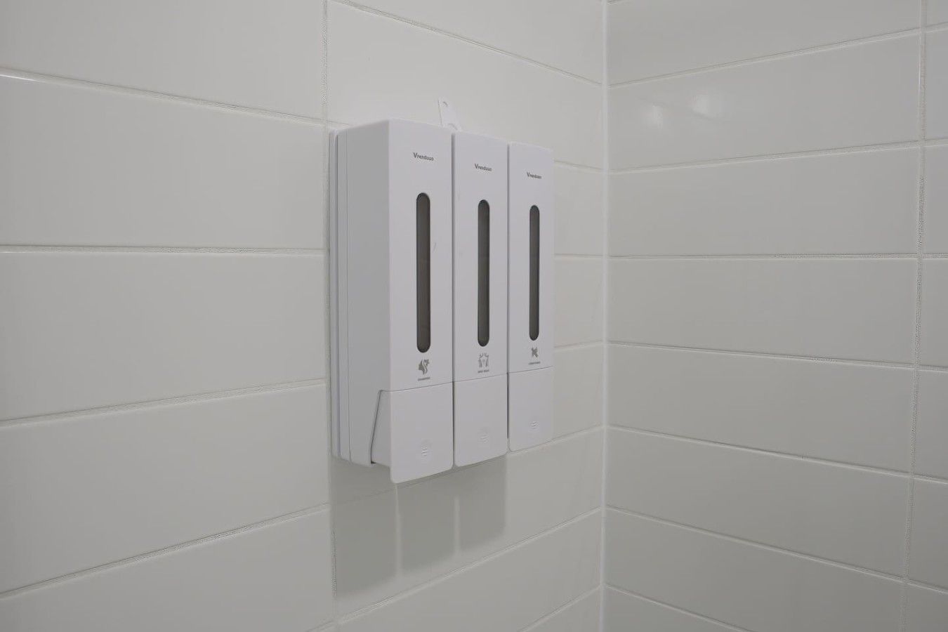 A wall-mounted hand sanitizer dispenser unit with a white design, mounted on a white tiled wall.
