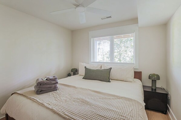 A bedroom with a bed, a window, a ceiling fan, and a bedside table with a lamp.