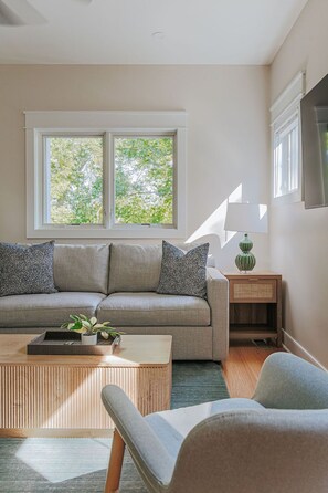 A living room with a sofa, a coffee table, and a side table with a lamp. There are windows with a view of trees. - Riverbluff Nest (Charlottesville)