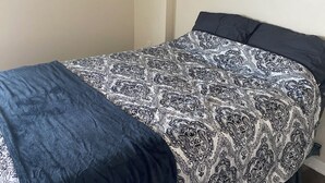 Ein Einzelbett mit einem gemusterten Überwurf und einer dunkelblauen Decke in einem Zimmer mit schlichten Wänden und Holzdielen.