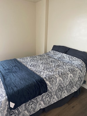 Een enkel bed met een bedovertrek met patroon en een donkerblauwe deken in een kamer met effen muren en houten vloeren.