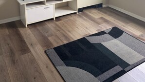 Ein Zimmer mit Fernseher auf einem weißen Schrank, einem Heizkörper, einem Fenster mit Jalousien und einem gemusterten Teppichboden.