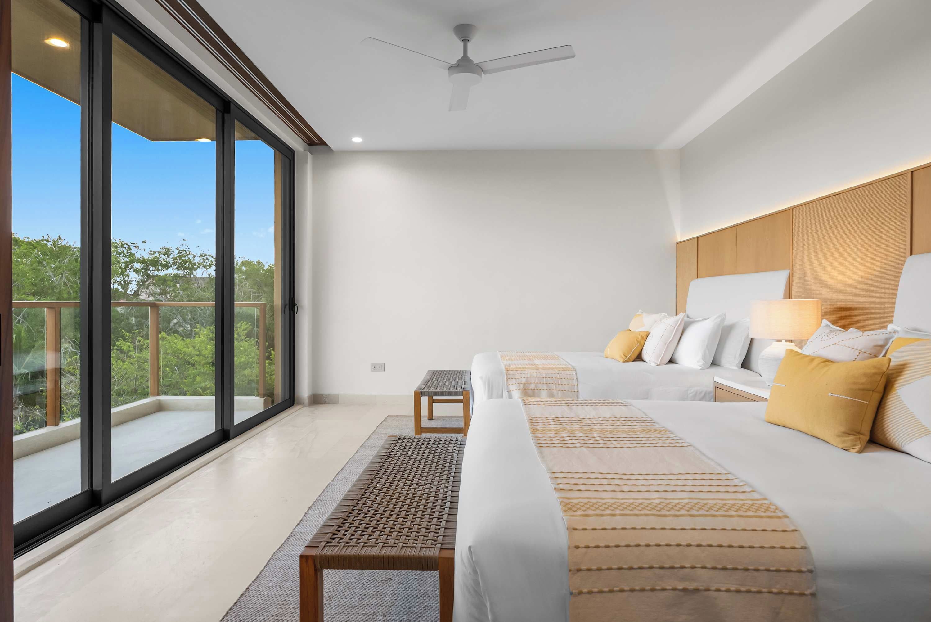 Ein modernes Schlafzimmer mit einer großen Schiebetür aus Glas, einem Deckenventilator, einem Bett mit weißer Bettwäsche und gelben Kissen sowie einem kleinen Holztisch.