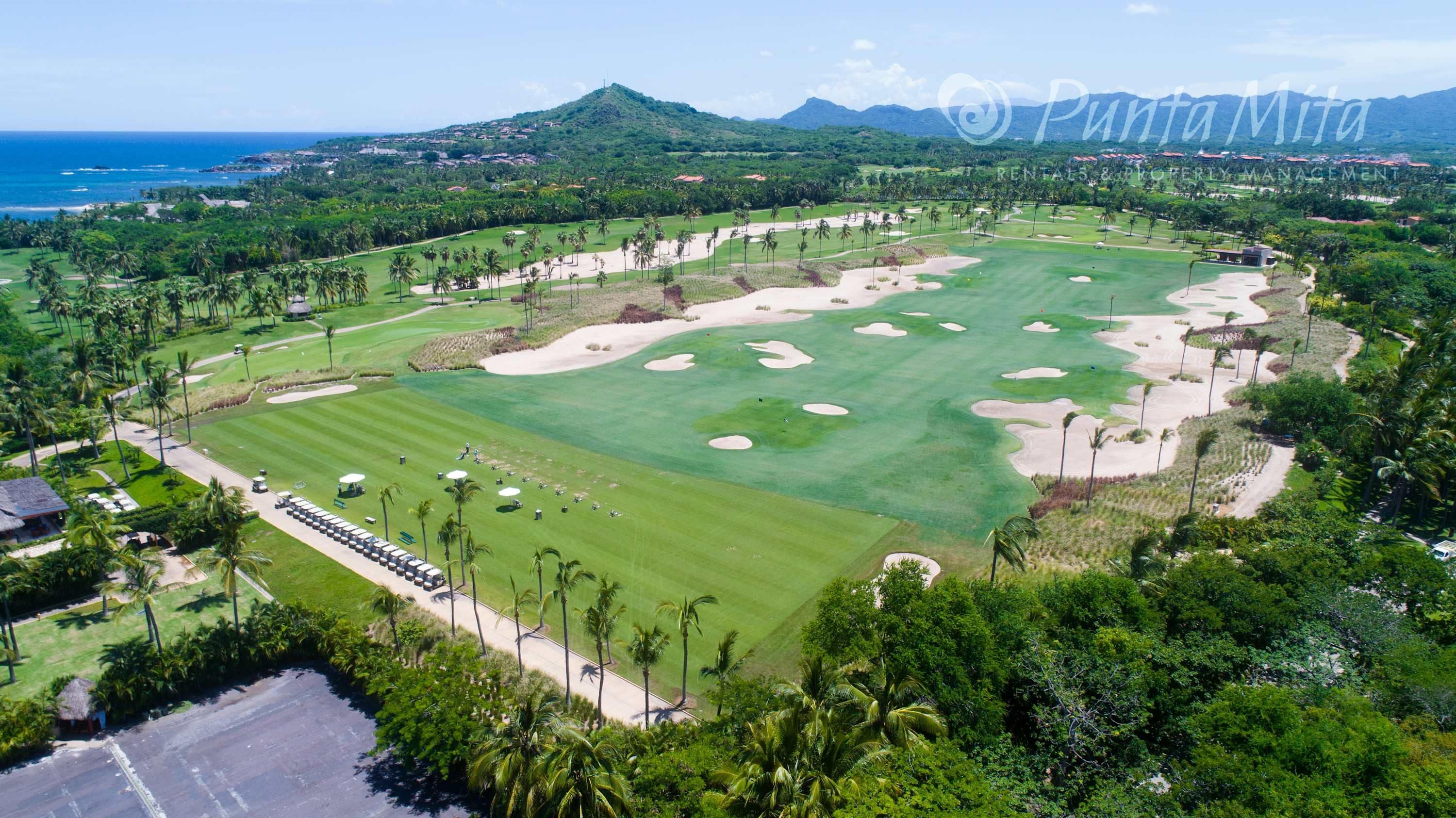An aerial view of a golf course with a coastal backdrop, surrounded by lush greenery and palm trees.