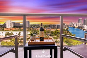 A balcony with a table holding a bottle of wine and two glasses, overlooking a cityscape with a river. - Stylish 1BR APT w/ Beach Access, Resort Amenities (Hallandale Beach)