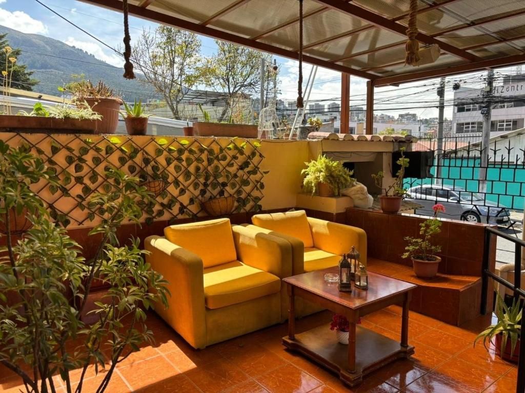 Un espacio de descanso al aire libre con techo, que incluye un sofá amarillo y una mesa de madera, rodeado de plantas en macetas y una pared decorativa.