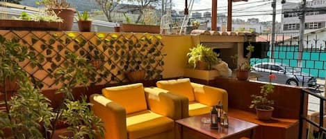 A covered outdoor seating area with a yellow sofa and a wooden table, surrounded by potted plants and a decorative wall.