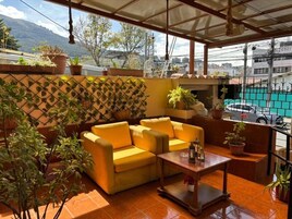 Área de estar coberta ao ar livre com um sofá amarelo e uma mesa de madeira, cercada por plantas em vasos e uma parede decorativa.