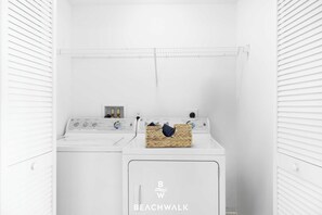 A laundry room with a front-loading washer and dryer, a basket of laundry, and a shelf with towels. - Bright Modern APT 1BR| Pool - Lounge chairs (Hallandale Beach)