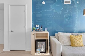 A room with a blue wall featuring a nautical chart, a wooden shelf with bottled water, a small refrigerator, and a grey sofa with a yellow pillow. - Bright Modern APT 1BR| Pool - Lounge chairs (Hallandale Beach)