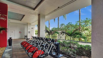 A covered outdoor area with a row of stationary bikes for rent, a pathway leading to a door marked 'PACKAGE ROOM,' and a view of palm trees and greenery.