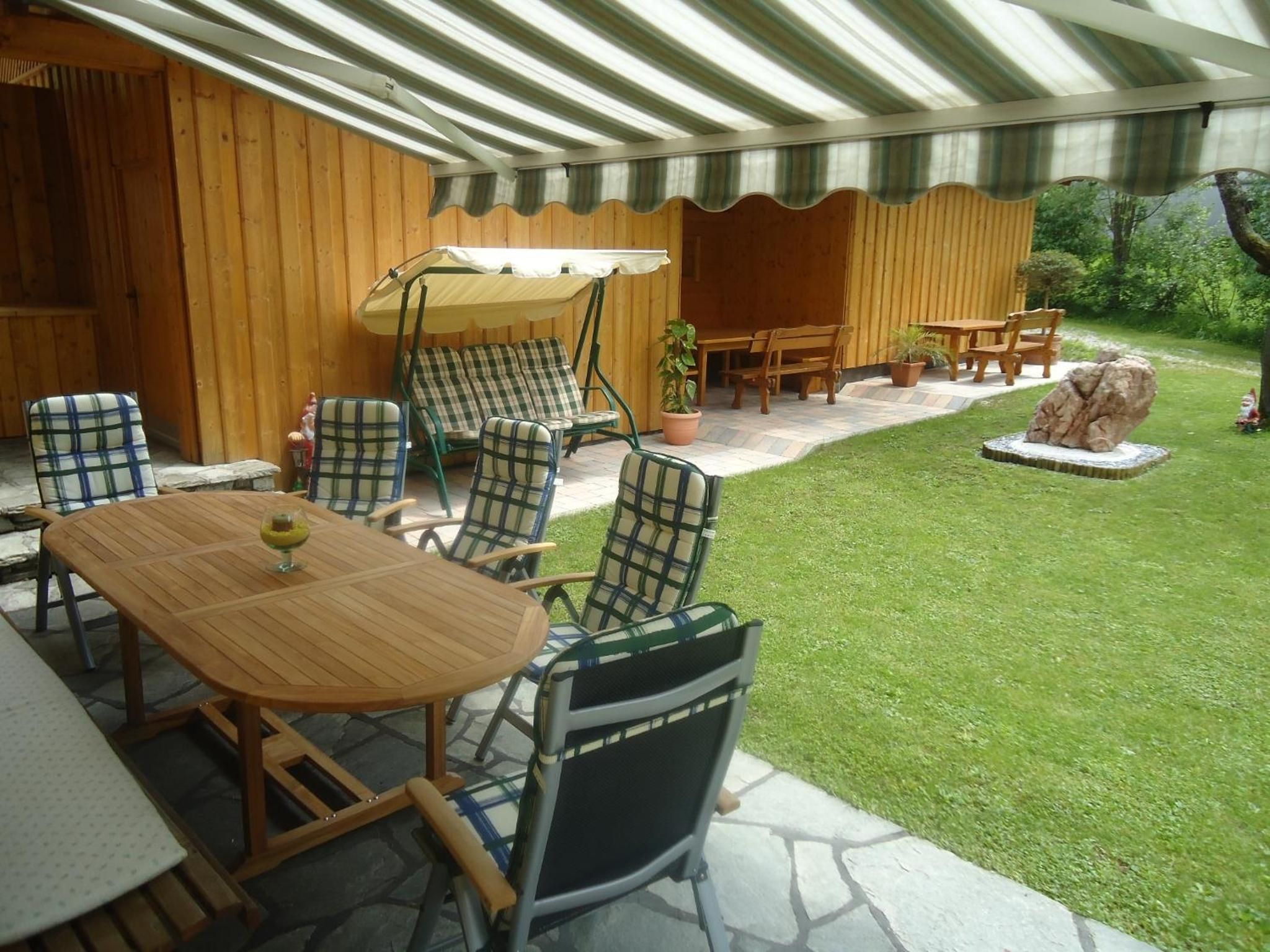 Un patio exterior con mesa y sillas de madera, un área de estar cubierta y un césped bien cuidado.