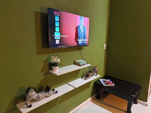 Habitación con un televisor de pared mostrando una foto de perfil y un programa titulado 'EL MENTALISTA'. Hay estantes blancos con objetos decorativos y una mesa negra con una revista.