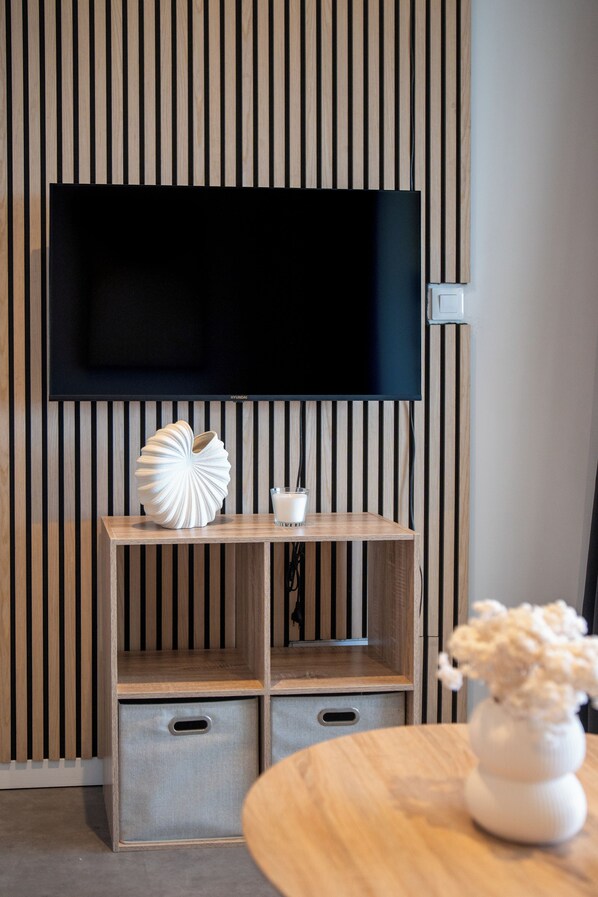 Un salon moderne avec un téléviseur à écran plat fixé à un mur composé de lattes de bois, un meuble TV en bois agrémenté d’un objet décoratif et d’un verre de lait, et une table ronde en bois sur laquelle est posé un vase de fleurs.