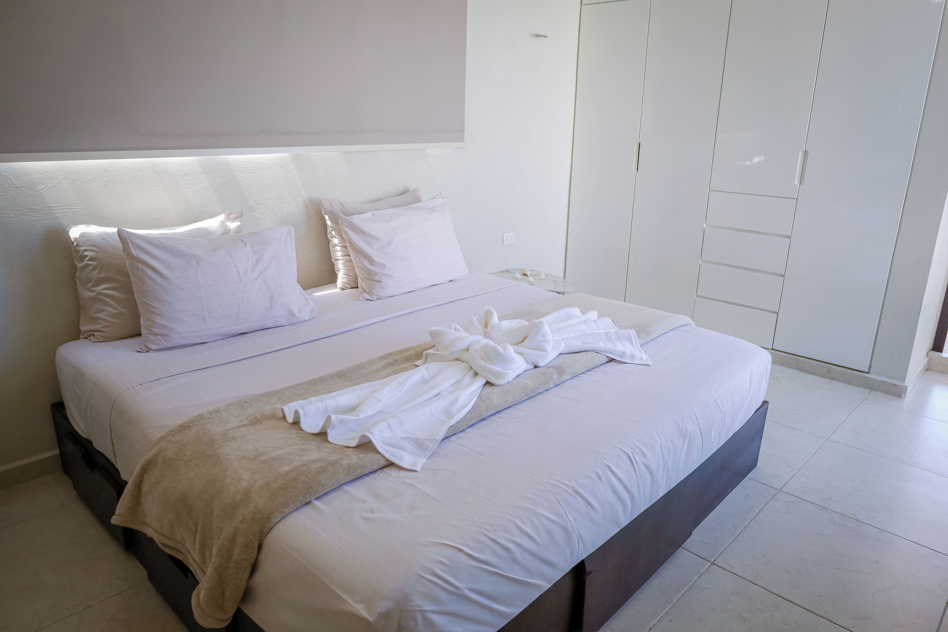 A neatly made bed with white linens and a beige throw blanket, a bedside table with a glass top, and a white wardrobe with sliding doors.