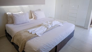 Cama bem arrumada com lençóis brancos e manta bege, mesa de cabeceira com tampo de vidro e guarda-roupa branco com portas de correr.