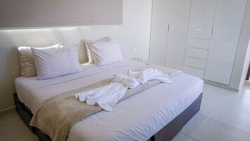 A neatly made bed with white linens and a beige throw blanket, a bedside table with a glass top, and a white wardrobe with sliding doors.
