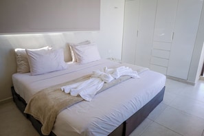 Ein ordentlich bezogenes Bett mit weißen Leinen und einer beigen Tagesdecke, ein Nachttisch mit Glasplatte und ein weißer Kleiderschrank mit Schiebetüren.