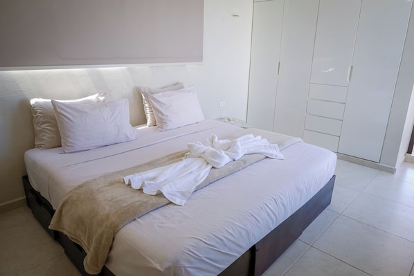 Ein ordentlich bezogenes Bett mit weißen Leinen und einer beigen Tagesdecke, ein Nachttisch mit Glasplatte und ein weißer Kleiderschrank mit Schiebetüren.