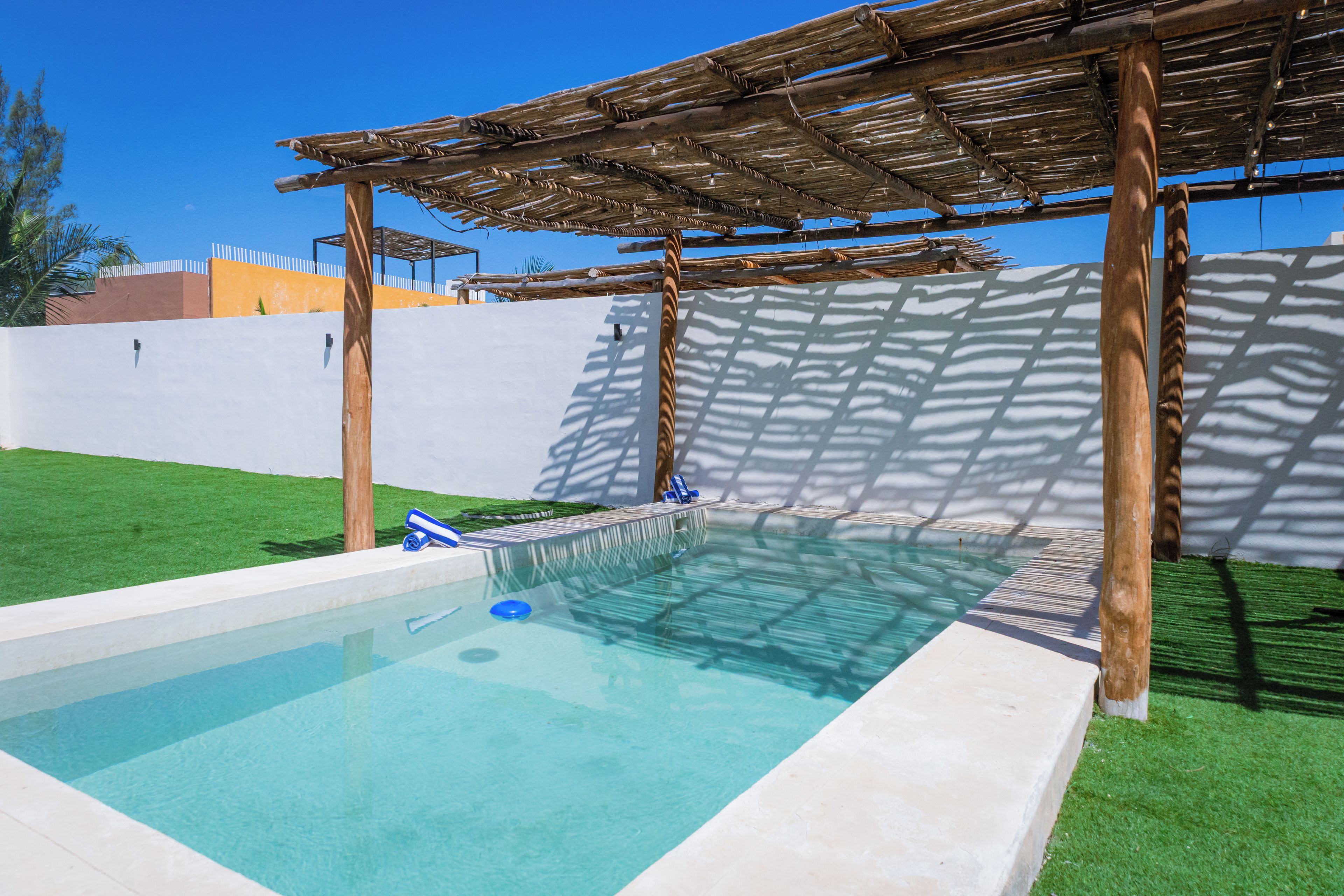 Una piscina con una pérgola de madera sobre ella, rodeada por un muro blanco y un césped verde.