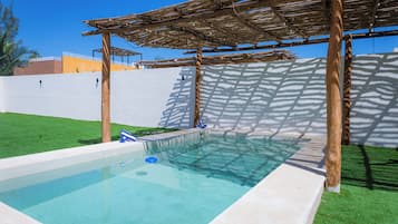 Una piscina con una pérgola de madera sobre ella, rodeada por un muro blanco y un césped verde.